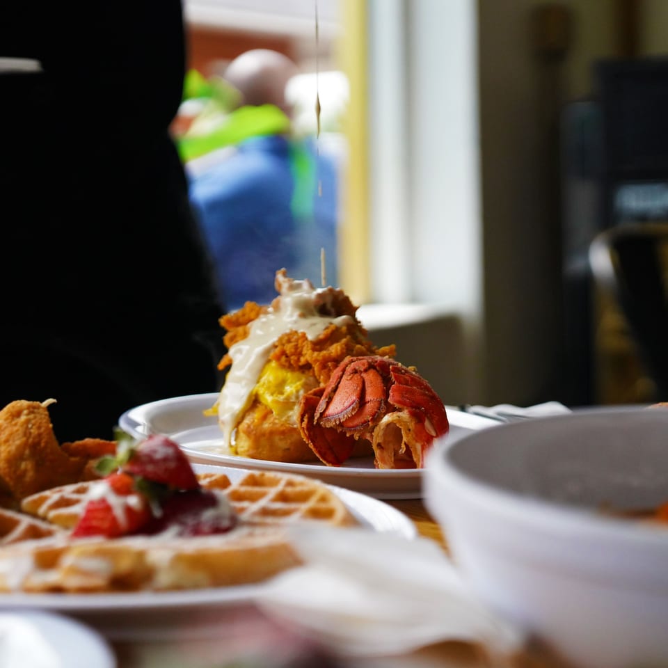 Syrup pouring over fried chicken, lobster, and scrambled eggs, with waffles and strawberries in the foreground.
