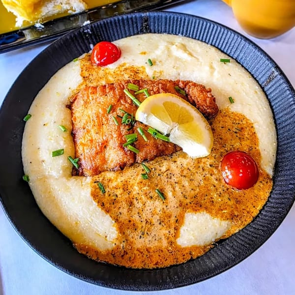 A black bowl of creamy grits topped with fried fish, a lemon slice, and cherry tomatoes.