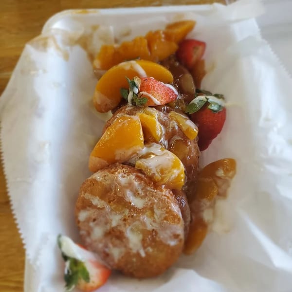 Golden-brown fritters topped with peaches, strawberries, and a sweet glaze, served in a white paper container.
