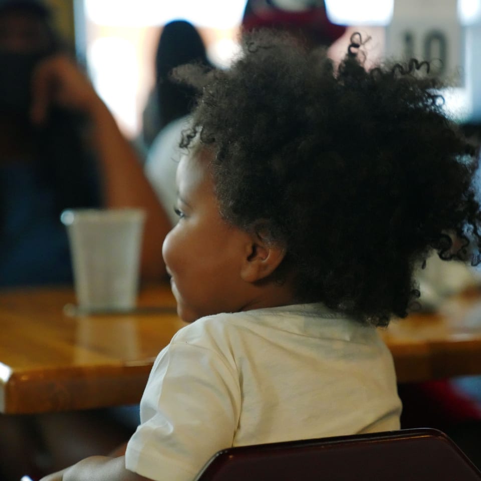 A young child with curly hair smiles, looking right, seated at a table in The Real Milk & Honey.