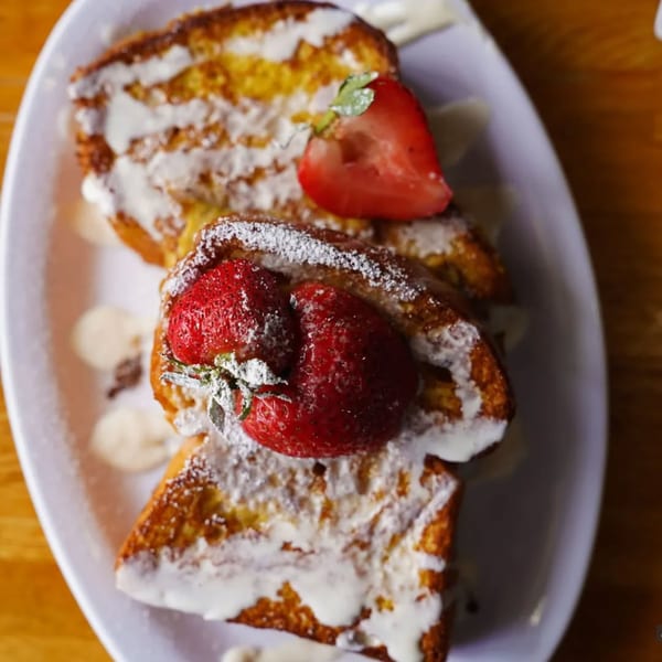 Golden French toast topped with powdered sugar, white icing, and fresh strawberries on a white plate.