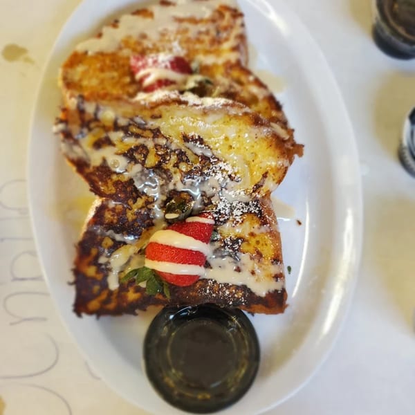 Golden French toast drizzled with white icing, powdered sugar, strawberries, and a side of syrup.