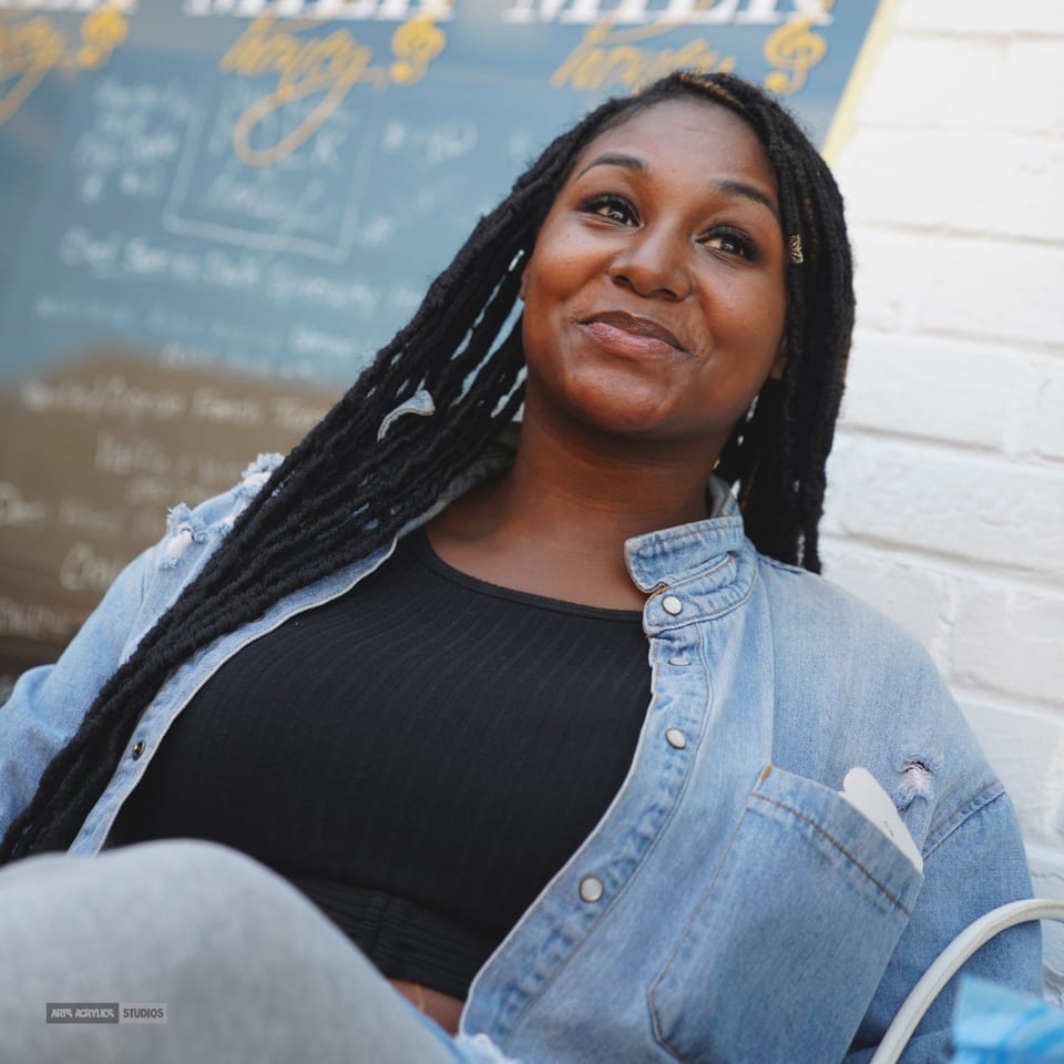 A smiling woman with long dreadlocks and a denim jacket looks up, with a menu board behind her.