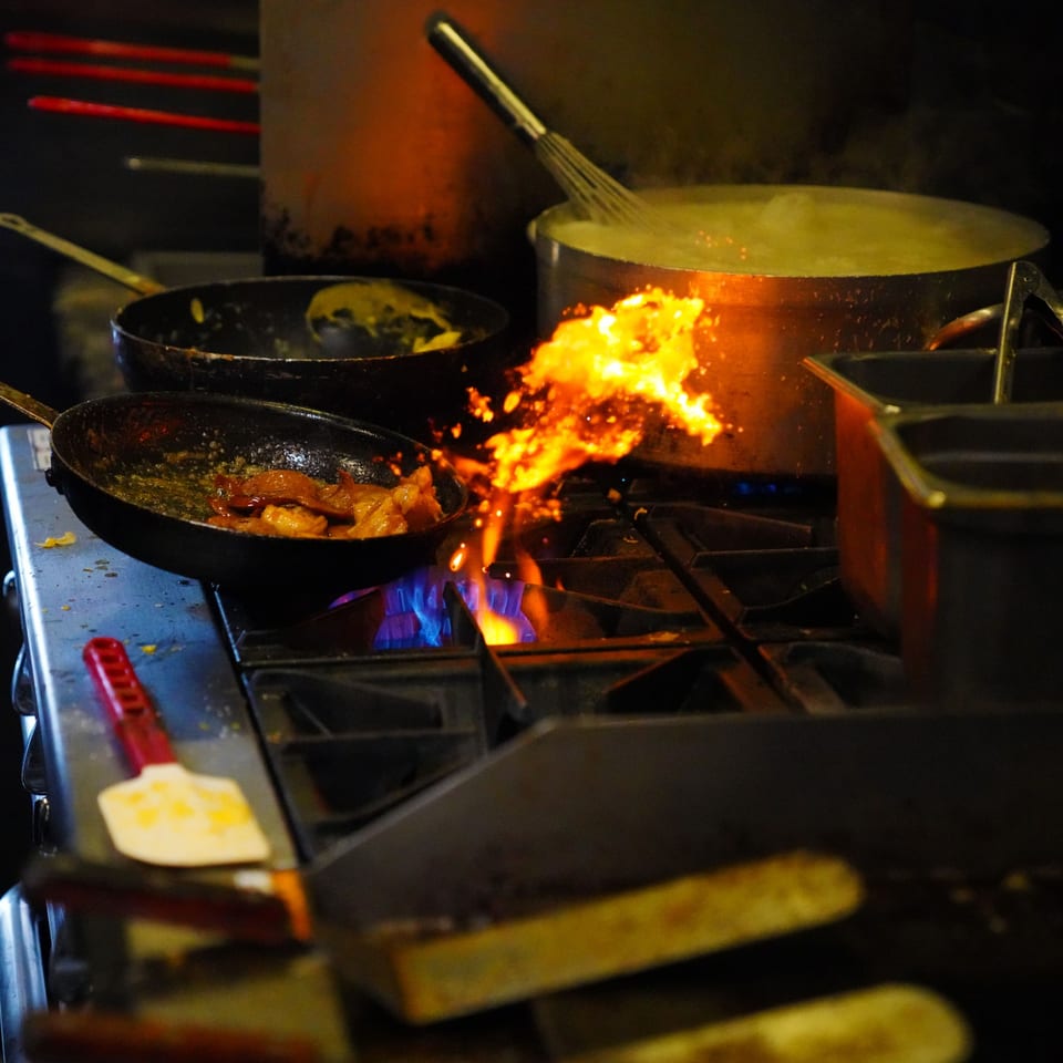 A chef flambés food in a pan on a stove, with another pot boiling beside it.