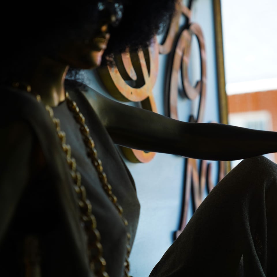 Mannequin with afro and gold chain necklace, against a decorative metal screen.