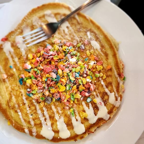 Golden pancakes topped with white icing, colorful Fruity Pebbles cereal, and a fork on a white plate.