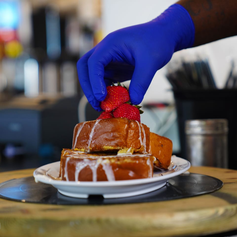A gloved hand places fresh strawberries on a stack of syrup-drizzled French toast at The Real Milk & Honey.