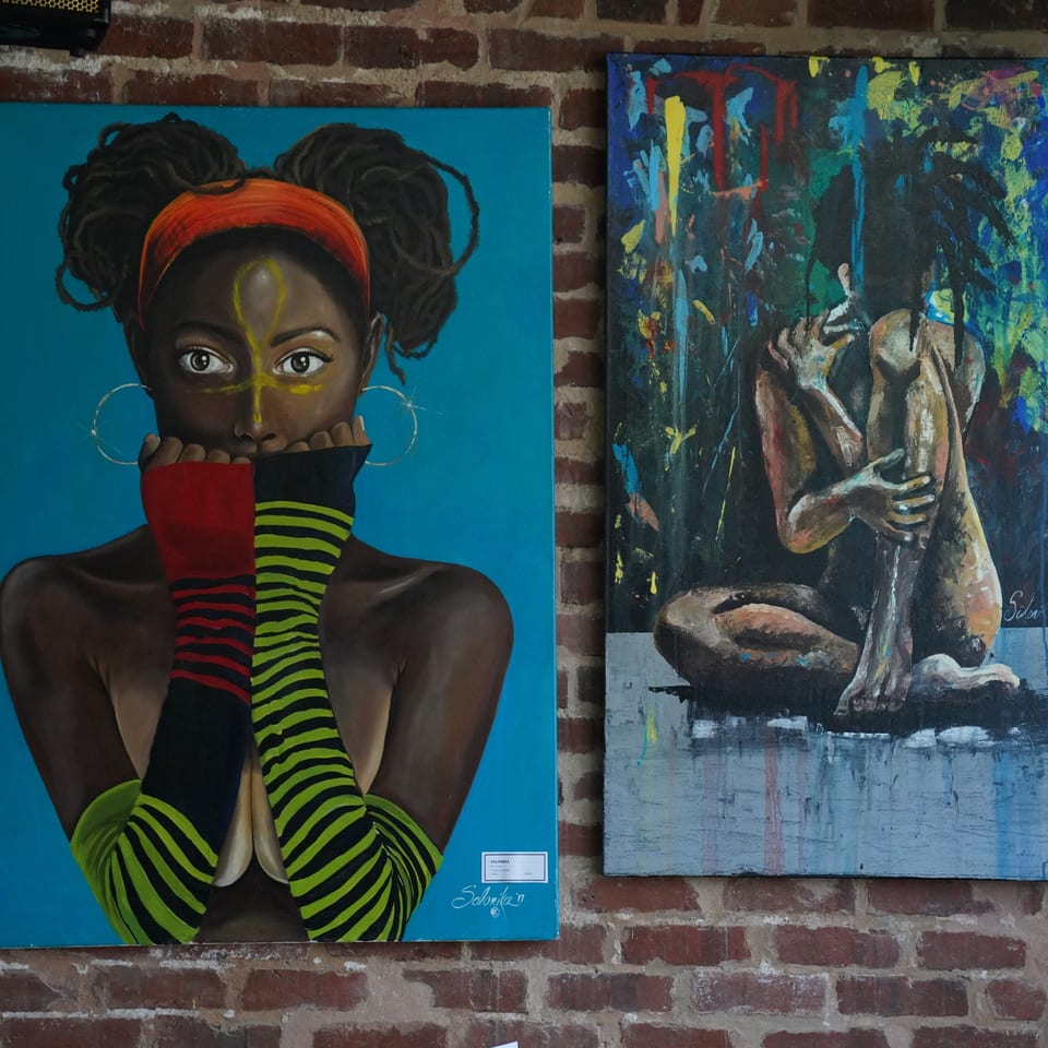 Two vibrant, abstract paintings of figures, one with dreadlocks, hanging on a brick wall.