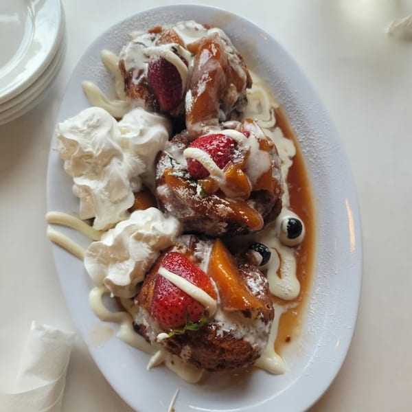 A plate of golden-brown French toast bites topped with strawberries, peaches, whipped cream, and syrup.